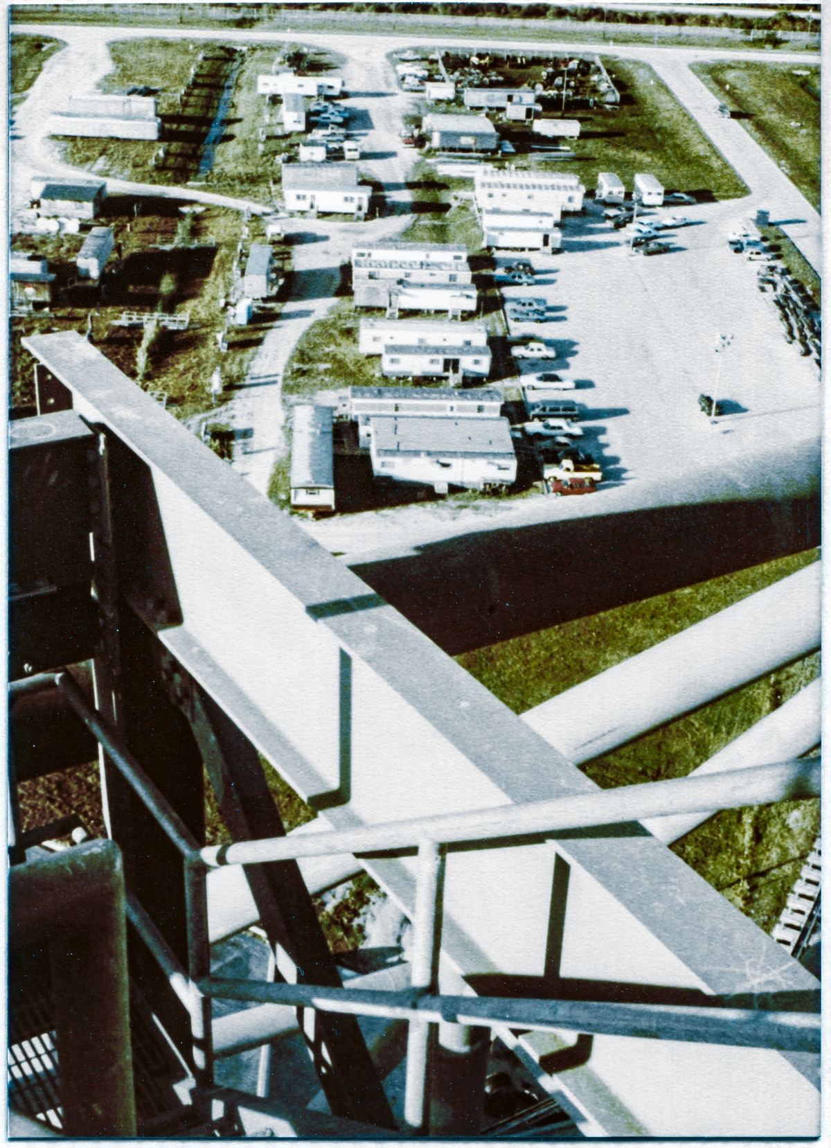 Image 028. From the roof of the Rotating Service Structure at Launch Complex 39-B, Kennedy Space Center, Florida, at an elevation of over 200 feet above ground level, you are looking across part of the top perimeter framing of Stair Tower 4, with the large structural pipes of the RSS Primary Framing of Column Lines 6 and 7 beyond and below that, to the area where NASA engineering and the contractor’s jobsite administrative field trailers were located around an asphalt parking lot. Behind and beyond those trailers, in an area of bare grass and dirt roads, a variety of contractor’s tool, equipment, and storage trailers can also be seen. Photo by James MacLaren.